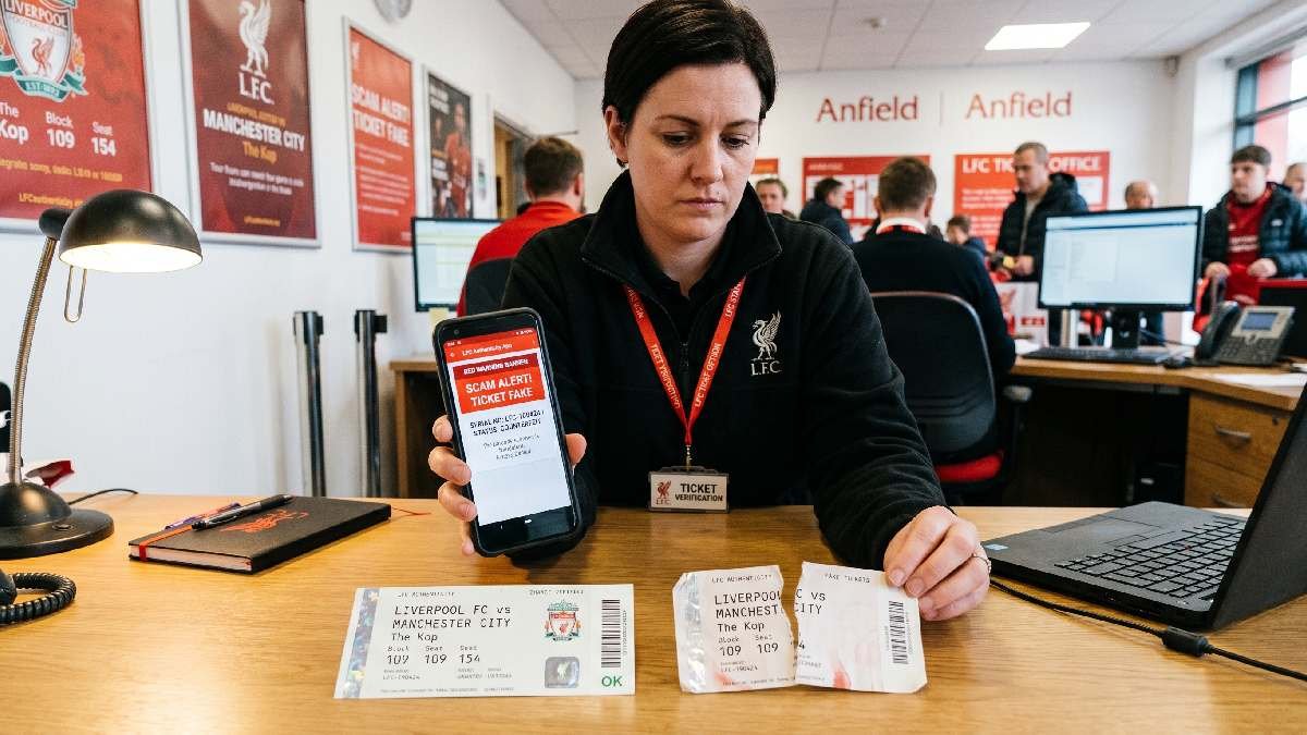 Person checking Liverpool FC ticket authenticity on smartphone with scam alert and fake ticket visible on desk.