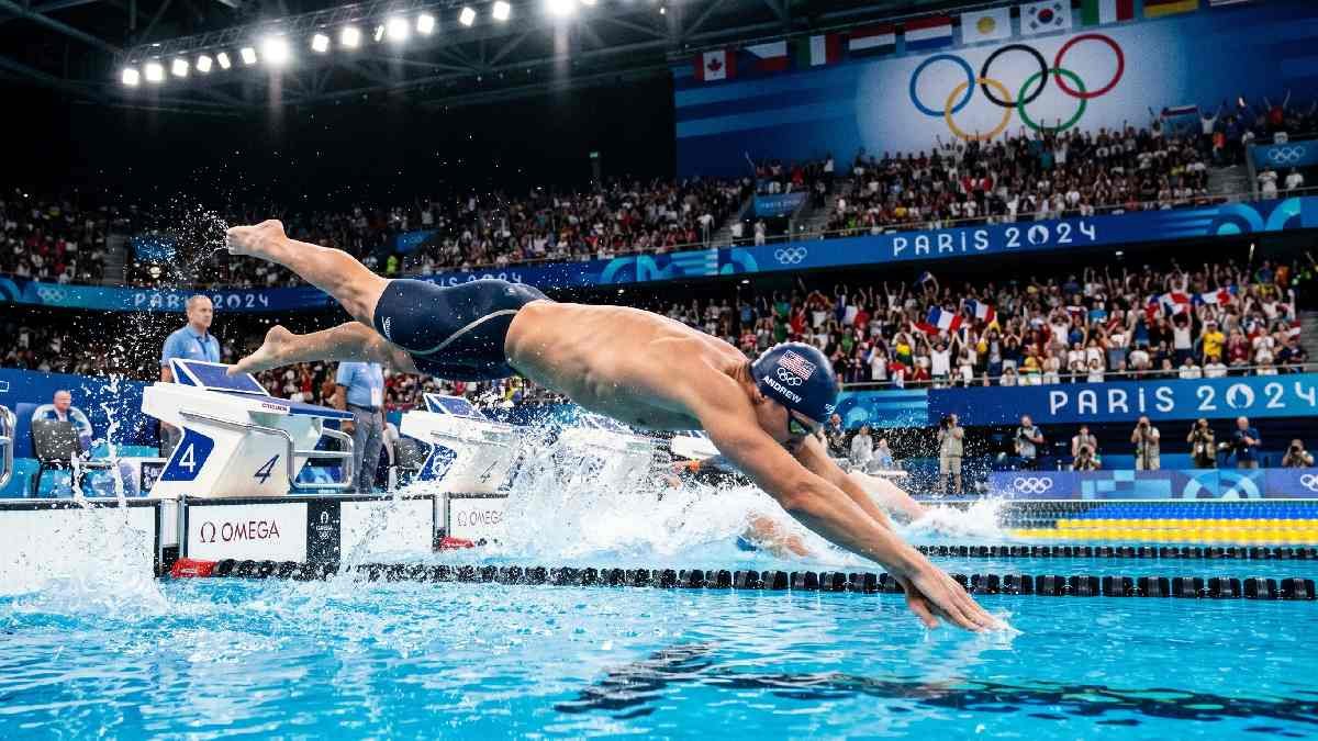 Olympic swimmer diving into a competition pool with Olympic rings banner and packed crowd in background.