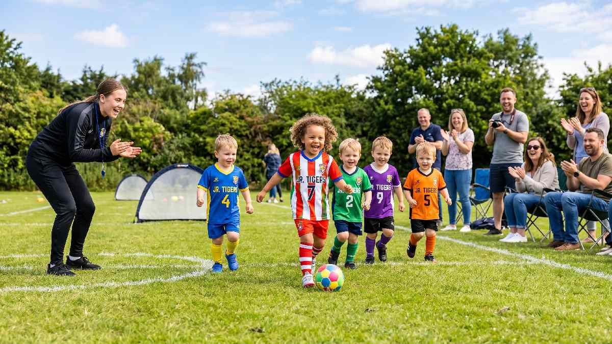 Happy toddlers in colorful football kits kicking a ball on grass field with coach and smiling parents watching.